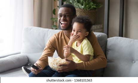 Family sitting on couch in living room spending weekend together at home, african father holding on lap little son using remote control watching funny movie comedy laughing having fun eating popcorn