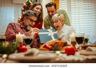 Family is sitting at their christmas dinner table, enjoying their meal and each other's company while opening a present - Powered by Shutterstock