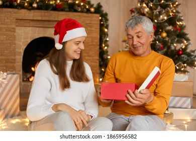 Family Sitting With Their Grandfather Celebrating Christmas In Cozy House, Woman In Santa Claus Hat Giving New Year Present, Happy Old Man Opening Gift With Pleasant Smile.