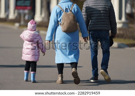Family with small child walk on the spring city street. Mother will lead their daughter by the hand.  She put on pink clothes, rubber boots.Time for leisure and walking with family. Low angle view. 