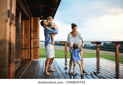 Family with small children playing in rain on patio by wooden cabin, holiday in nature concept.