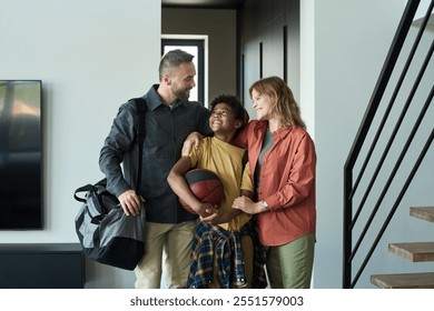 Family smiling and greeting each other at the doorway, with child holding basketball and looking happy. Modern interior with flat screen TV and staircase visible in background - Powered by Shutterstock