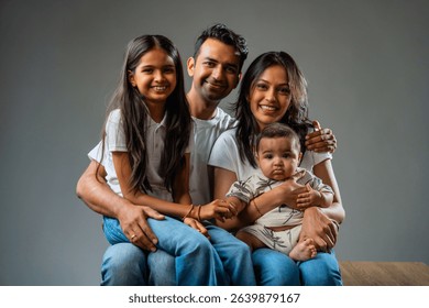 Family smiling looking at camera while sitting together, Indian parents with infant and daughter indoors on gray background, isolated and cheerful, expressing love, happiness, bonding and connection