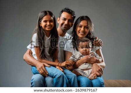 Family smiling looking at camera while sitting together, Indian parents with infant and daughter indoors on gray background, isolated and cheerful, expressing love, happiness, bonding and connection