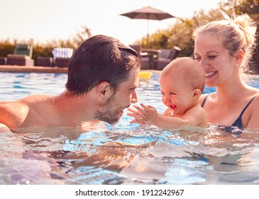 Family With Son And Baby Daughter Having Fun On Summer Vacation Splashing In Outdoor Swimming Pool