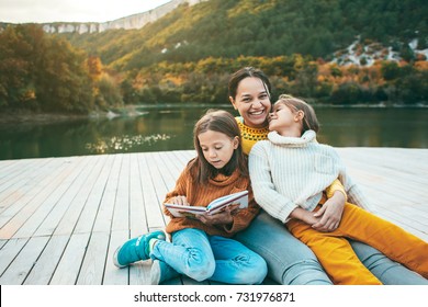 Family Spending Time Together By The Lake In Autumn. Mom With Her Daughters Reading Book Near The Pond. Fall Weekend In The Open Air.