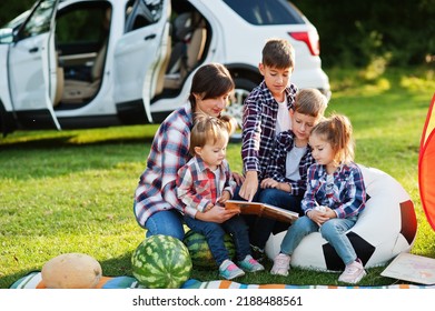 Family Spending Time Together. Mother Reading Book Outdoor With Kids Against Their Suv Car.