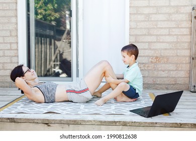 Family Sport Activity. Middle Age Caucasian Mother With Toddler Doing Sit-ups Exercices On Backyard. Distant Remote Online Fitness Workout With Laptop Outdoors. Parent Training With Child Boy Kid. 