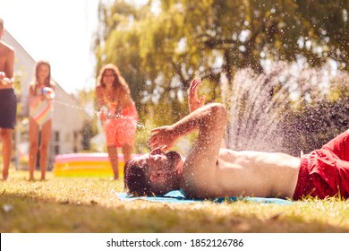 Family Squirting Father With Water Pistols In Summer Garden As He Sunbathes