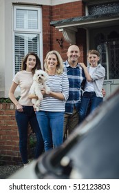 Family Standing Outside Their Home Together With Their Pet Dog, Smiling For The Camera. 