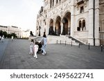 Family strolls with children and stroller near historic building on an open plaza. Urban sightseeing and family outing.