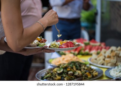 Family Style Self Serve Buffet Showcasing Colorful Greek Food With Female Loading Plate In Focus And Remainder Of Buffet In Background In Soft Focus