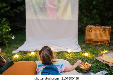 Family Summer Outdoor Movie Night. Girl Lying On Blanket, Eating Homemade Popcorn And Watching Film On  DIY  Screen With From Projector. Summer Outdoor Weekend Activities With Kids. Open Air Cinema