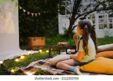 Family summer outdoor movie night. Girl sitting on blanket and pillows, eating homemade popcorn and watching film on DIY screen with from projector. Summer outdoor weekend activities with kids. - Shutterstock ID 2112353705