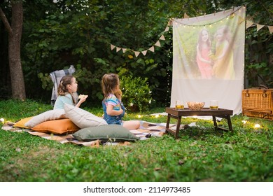 Family Summer Outdoor Movie Night. Girls Sitting On Blanket And Pillows, Eating Homemade Popcorn And Watching Film On DIY Screen With From Projector. Summer Outdoor Weekend Activities With Kids.