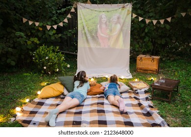 Family Summer Outdoor Movie Night. Girls Lying On Blanket And Pillows, Eating Homemade Popcorn And Watching Film On DIY Screen With From Projector. Summer Outdoor Weekend Activities With Kids.