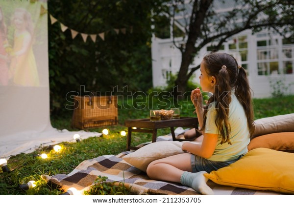 Family summer outdoor movie night. Girl sitting on\
blanket and pillows, eating homemade popcorn and watching film on\
DIY screen with from projector. Summer outdoor weekend activities\
with kids.