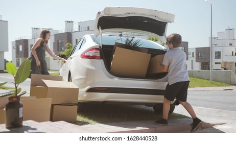 Family Summer Roadtrip To The Seaside. Two Cute Siblings Sister And Brother Loading A Heavy Suitcase In Trunk Standing Near The Car In The Courtyard.