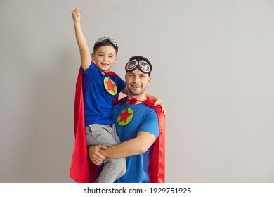 Family in superhero costumes. Father holds his son in his arms standing on a gray background. Dad and boy in red cloaks, pilot glasses and with stars on his chest celebrate Father's Day. Banner. - Shutterstock ID 1929751925