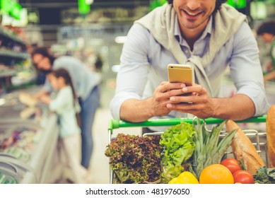 Family In The Supermarket. Cropped Image Of Dad Leaning On Shopping Cart, Using A Mobile Phone And Smiling, In The Background His Wife And Daughter Are Choosing Food