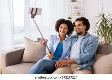 Family, Technology And People Concept - Happy Couple With Smartphone And Selfie Stick Taking Picture At Home