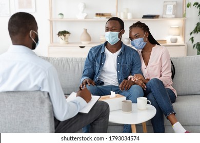 Family Therapy During Pandemic. Happy Black Couple In Protective Masks Sitting On Couch At Counselor's Office, Listening Advices, Selective Focus