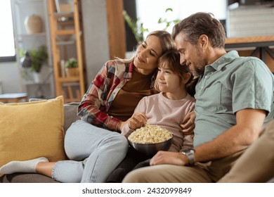 A family of three is comfortably nestled on a couch, their faces reflecting excitement and attentiveness as they share a bowl of popcorn during a suspenseful movie night - Powered by Shutterstock