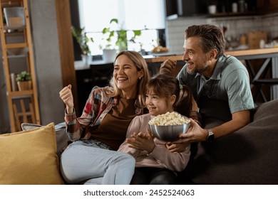 A family of three is comfortably nestled on a couch, their faces reflecting excitement and attentiveness as they share a bowl of popcorn during a suspenseful movie night - Powered by Shutterstock