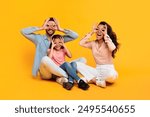 A family of three, a father, mother, and young daughter, are sitting on a yellow background. They are all making silly faces with their fingers over their eyes. They are having fun together.