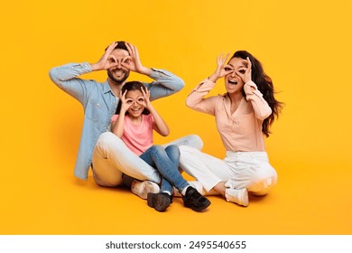 A family of three, a father, mother, and young daughter, are sitting on a yellow background. They are all making silly faces with their fingers over their eyes. They are having fun together. - Powered by Shutterstock