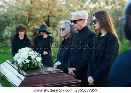 Family of three grieving people in sunglasses and mourning clothes standing in front of coffin with white flowers on lid during funeral service