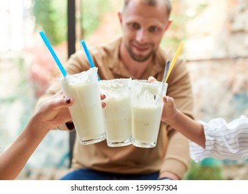 Family Of Three Having Drinks, Clinking Glasses With Milkshakes. Smiling Father Is Blurred On Background. Focus Is On Milkshakes. Concept Of Restaurant, Drinks And Hospitality.