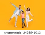 A family of three is posing for a photo in a studio. The father is striking a playful pose with his arm outstretched, while the mother and daughter are jumping and smiling brightly.