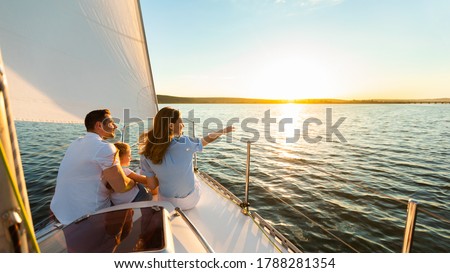 Family Of Three Sailing On Yacht Sitting On Sailboat Deck Looking At Sunset At Seaside. Dream Vacation Concept. Free Space