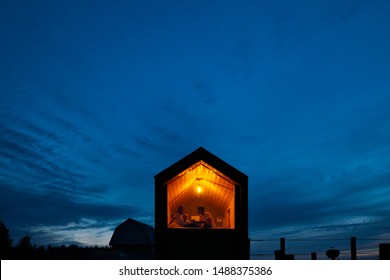 A family of three is sitting on a bed by the window, reading a book and talking. View of the outside of the house. Night blue sky. Orange warm light from an incandescent lamp. The concept of home
