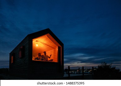 A Family Of Three Is Sitting On A Bed By The Window, Reading A Book And Talking. View Of The Outside Of The House. Night Blue Sky. Orange Warm Light From An Incandescent Lamp. The Concept Of Home