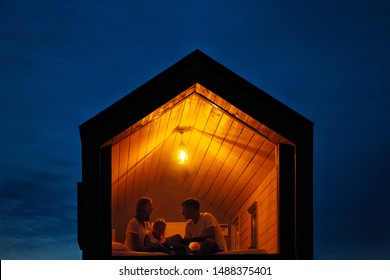 A Family Of Three Is Sitting On A Bed By The Window, Reading A Book And Talking. View Of The Outside Of The House. Night Blue Sky. Orange Warm Light From An Incandescent Lamp. The Concept Of Home