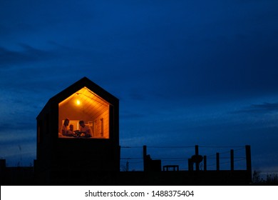 A Family Of Three Is Sitting On A Bed By The Window, Reading A Book And Talking. View Of The Outside Of The House. Night Blue Sky. Orange Warm Light From An Incandescent Lamp. The Concept Of Home