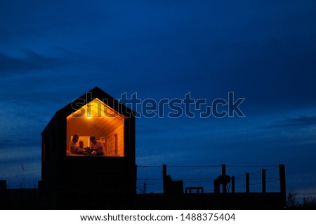 A family of three is sitting on a bed by the window, reading a book and talking. View of the outside of the house. Night blue sky. Orange warm light from an incandescent lamp. The concept of home