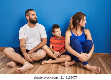 Family Of Three Sitting On The Floor At Home Looking To Side, Relax Profile Pose With Natural Face With Confident Smile. 