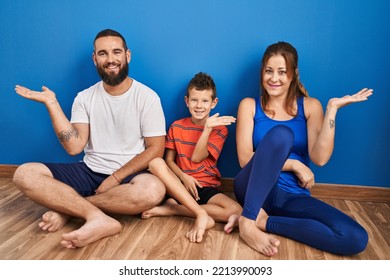 Family Of Three Sitting On The Floor At Home Smiling Cheerful Presenting And Pointing With Palm Of Hand Looking At The Camera. 