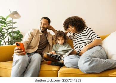 Family of three sitting relaxed on a yellow couch spending time together at home. The mother is engaging while the child reads a book, and the father is looking at his smartphone. - Powered by Shutterstock