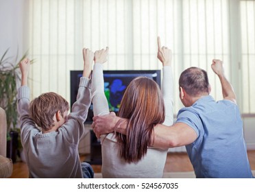 Family Of Three Watching Sports On TV And Cheering With Raised Arms. Rear View. 