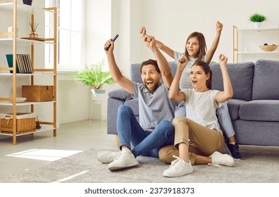 Family of three watching TV together. Family spending weekend at home and enjoying exciting football match on sports channel. Happy mom, dad and child celebrating goal, raising hands up and cheering - Powered by Shutterstock