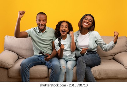 Family Time Activities. Happy Black Parents And Their Daughter Playing Video Games While Sitting On Sofa Over Yellow Background. Dad, Mother And Child Holding Joysticks, Winning Game