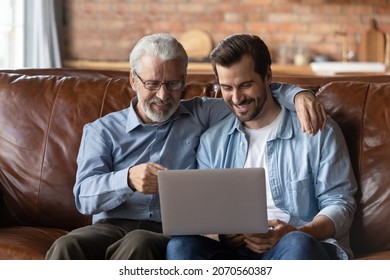 Family time with grown son. Elder and younger men relatives sit on sofa at home hug look on laptop screen discuss online news. Retired dad interested in pc talk ask adult child question about app work - Powered by Shutterstock