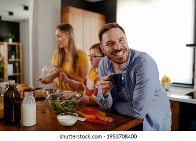 Family Time In Kitchen. Mom And Pretty Daughter Making A Lunch, While Smiling Dad Is Looking At Camera, Drinking A Coffee And Taking A Break From Cutting Vegetables. Focus On Man.