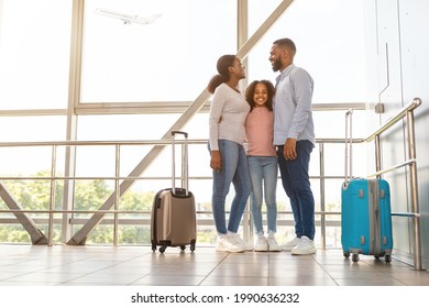 Family Travel Concept. Portrait Of Black Dad, Mum And Girl Standing At Departure Lounge With Suitcases Waiting For The Aircraft Arrival, Looking At Each Other And Hugging. Free Copy Space