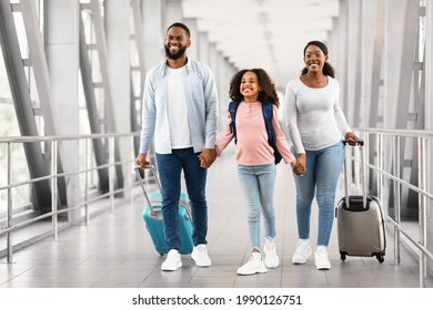 Family Travel Concept. Portrait of excited black dad, mum and girl walking at airport terminal corridor with suitcases waiting for the aircraft arrival, three people holding hands, free copy space - Shutterstock ID 1990126751
