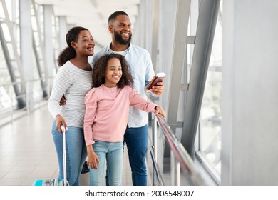 Family Travel Concept. Portrait Of Happy Black Man, Woman And Daughter Standing In Airport With Baggage And Looking At Aircraft Through Window. People Waiting For Air Plane Arrival In Departure Lounge